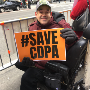 A man in a wheelchair holding a #SaveCDPA sign. He is wearing warm winter clothes and gloves.