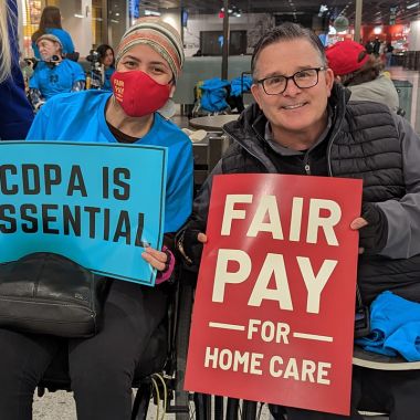 Womn and man, seated, holding "Fair Pay" and CDPA is Essential" signs